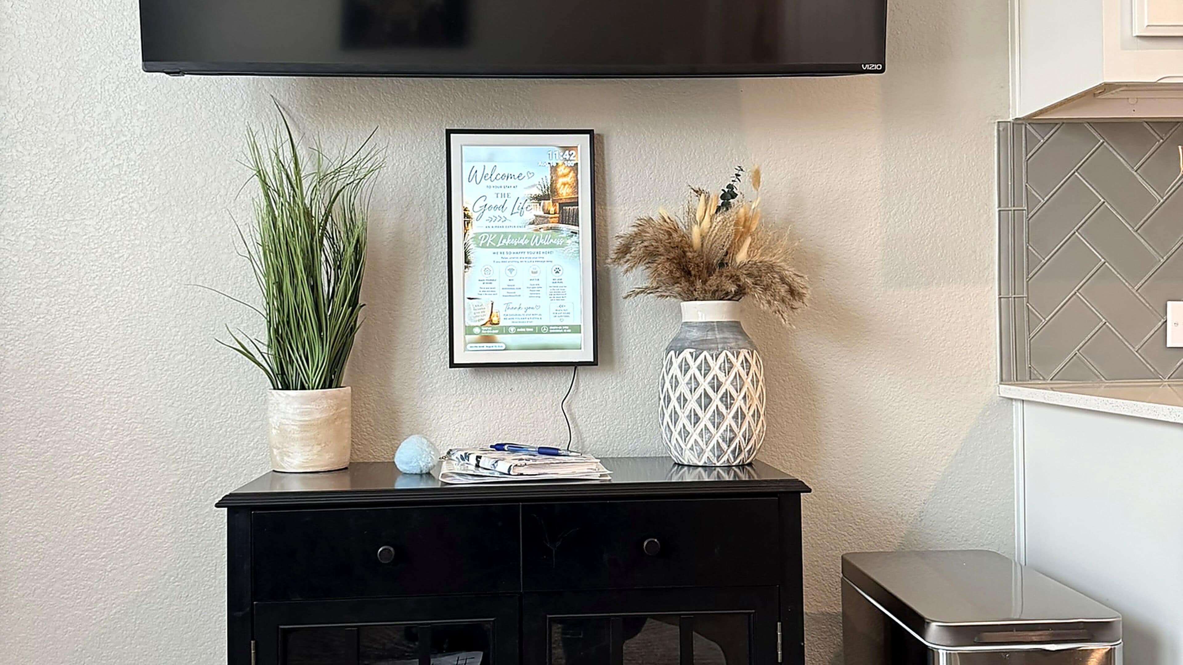 A black console table with a framed welcome sign, a potted plant, a decorative vase, and a small stack of papers.