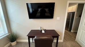 A room with a flat-screen TV mounted on the wall, a small table with a remote control, and a potted plant.