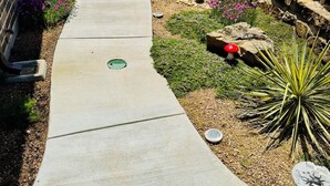 A concrete pathway leading to a wooden fence, with a brick wall on one side and a garden area with rocks, flowers, and a bee sculpture.