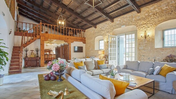 A spacious living area with a high ceiling, stone walls, and a wooden staircase leading to an upper level.