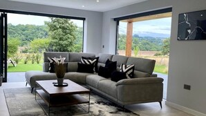 A modern living room with a grey sofa, black and white cushions, a wooden coffee table, and a large window offering a view of greenery and mountains. - Stunning modern home set in beautiful grounds. (Corston)