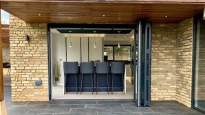 A modern outdoor patio with a wooden pergola, stone walls, and a seating area with chairs. - Stunning modern home set in beautiful grounds. (Corston)
