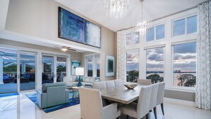 A spacious living room with a large dining table, comfortable seating, a ceiling fan, and large windows offering a view of the outdoors.