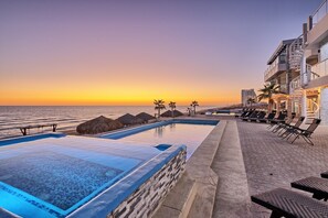A pool area with a view of the ocean at sunset, lounge chairs, and a building with a spiral staircase.