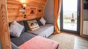 Et hyggeligt rum med trælofter, en grå sofa med puder, et pink plaid og udsigt over landskab gennem en glasdør.