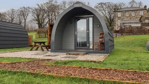 En lille, moderne, halvcirkelformet bygning med dør og vinduer, med teksten "LIVELONG VALLEY PODS", beliggende i et græsareal med et picnicbord og træbænke i nærheden.