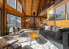 Ein großzügiger Wohnbereich mit grauem Sofa, Holztisch und einem Esstisch mit Stühlen. Große Fenster bieten Ausblick ins Freie.