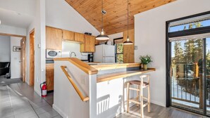 A modern kitchen with wooden cabinets, a central island with stools, pendant lighting, and a view of trees through a sliding glass door. - Secluded 3bed/Hot tub/Views (Fairplay)