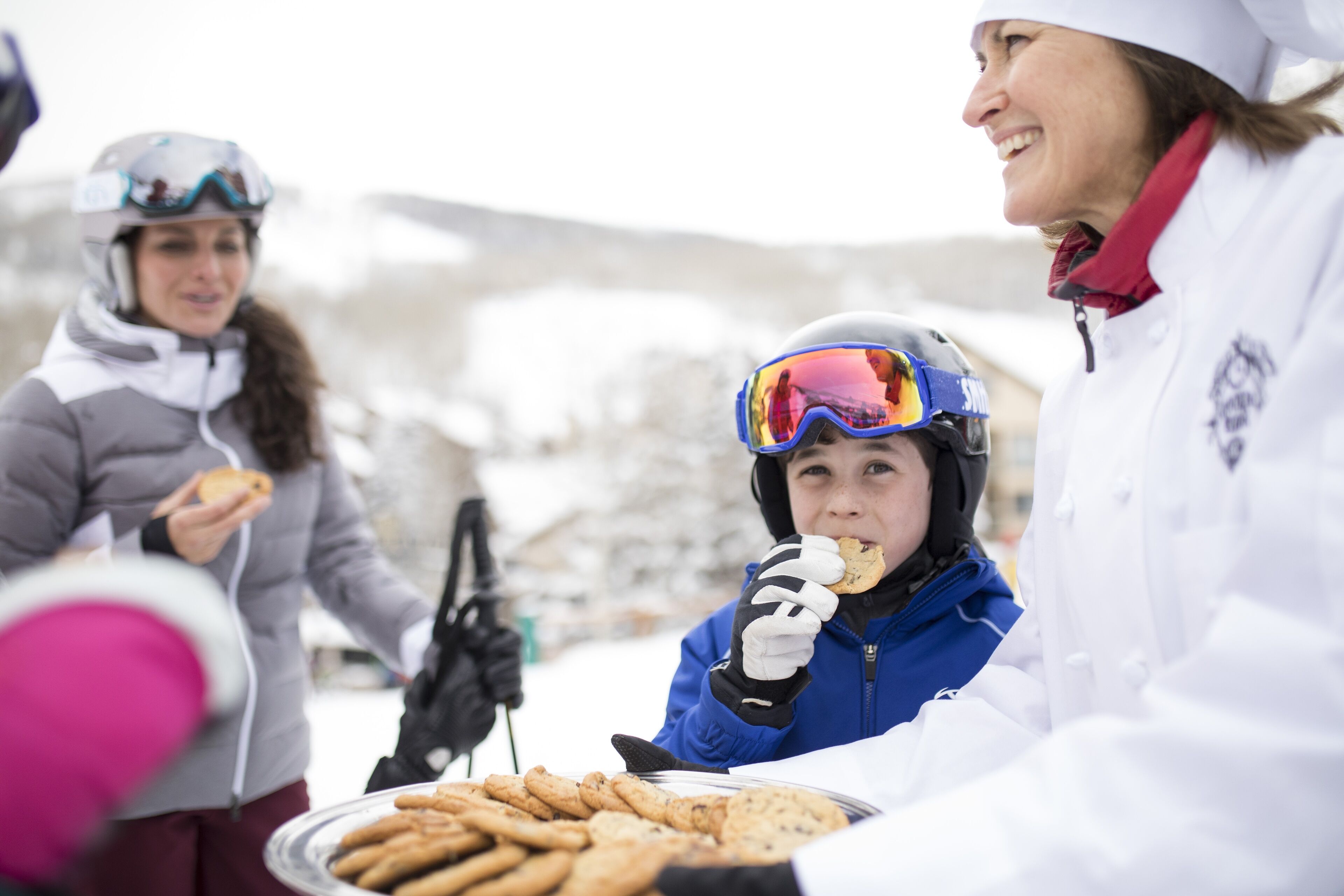 A group of people outdoors, some wearing winter gear, gathered around a table with food.