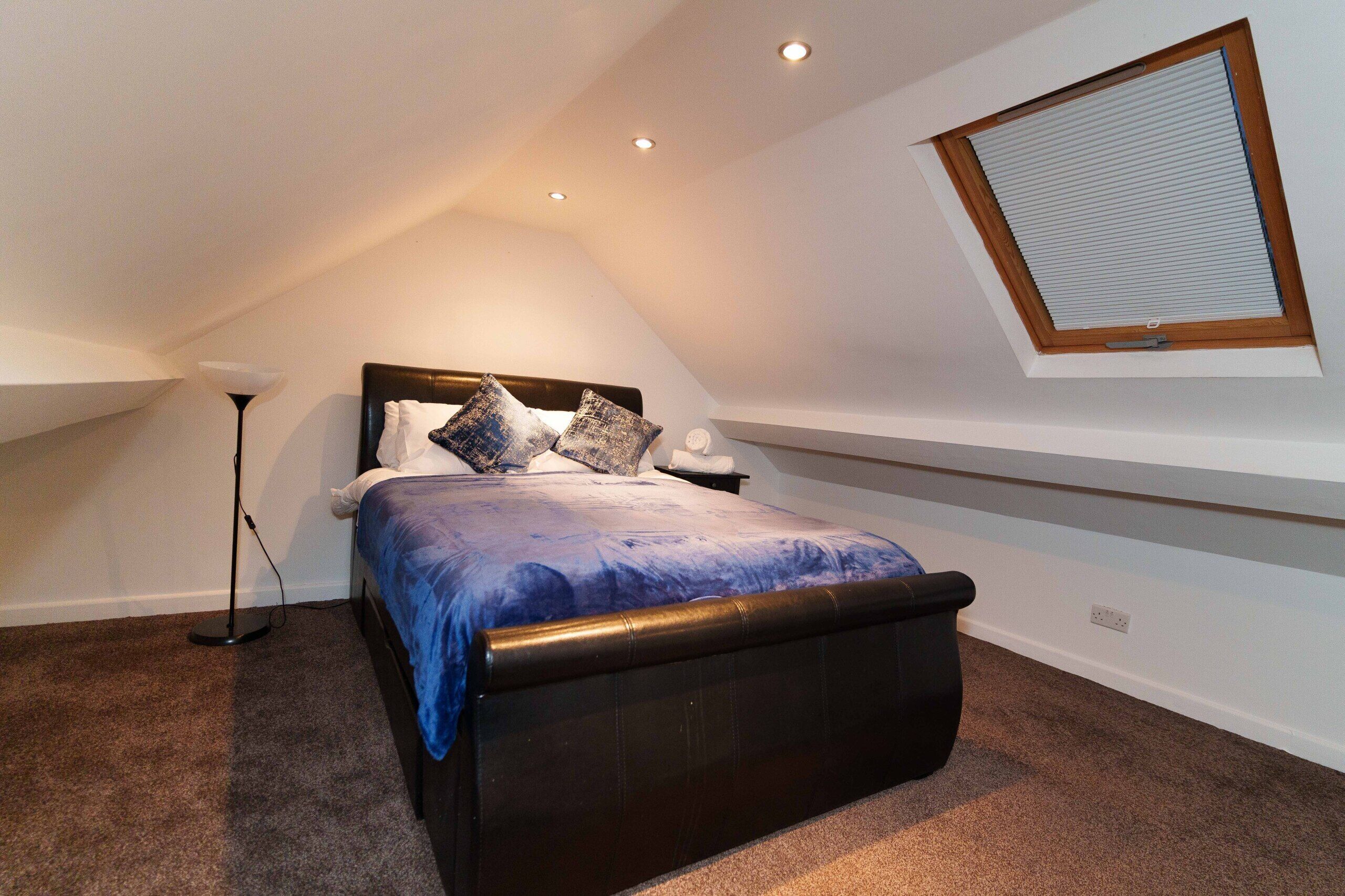 A bedroom with a sloped ceiling, a bed with a headboard, a bedside table, a lamp, and a window with blinds.