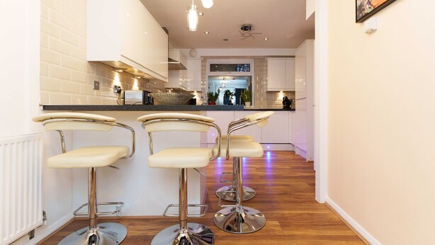 A modern kitchen with three bar stools, white cabinetry, and a wooden floor. - West Park House (Leeds)