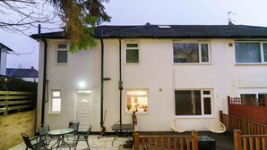 A two-story house with a patio area featuring a table and chairs, a wooden deck, and a small garden. - West Park House (Leeds)
