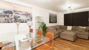 A modern living room with a glass dining table, brown leather chairs, a sectional sofa, and a cityscape painting on the wall. - West Park House (Leeds)