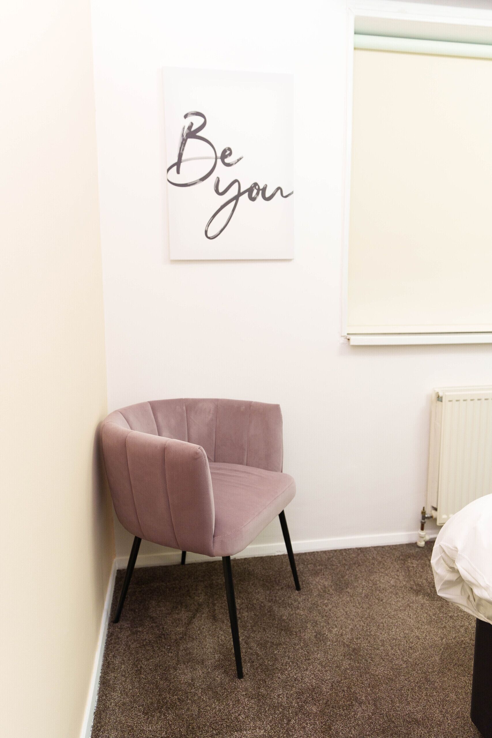 A room with a pink chair, a white radiator, and a wall with a framed sign that says "Be you".