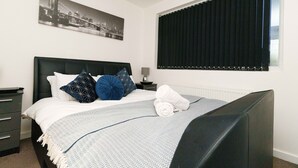 A neatly made bed with a patterned blanket, a cityscape artwork above it, and a window with blinds. - West Park House (Leeds)