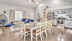 Une cuisine moderne dotée d’un four et d’un réfrigérateur intégrés, un coin repas avec une table en bois et des chaises blanches, et un salon avec un canapé bleu et un fauteuil suspendu.