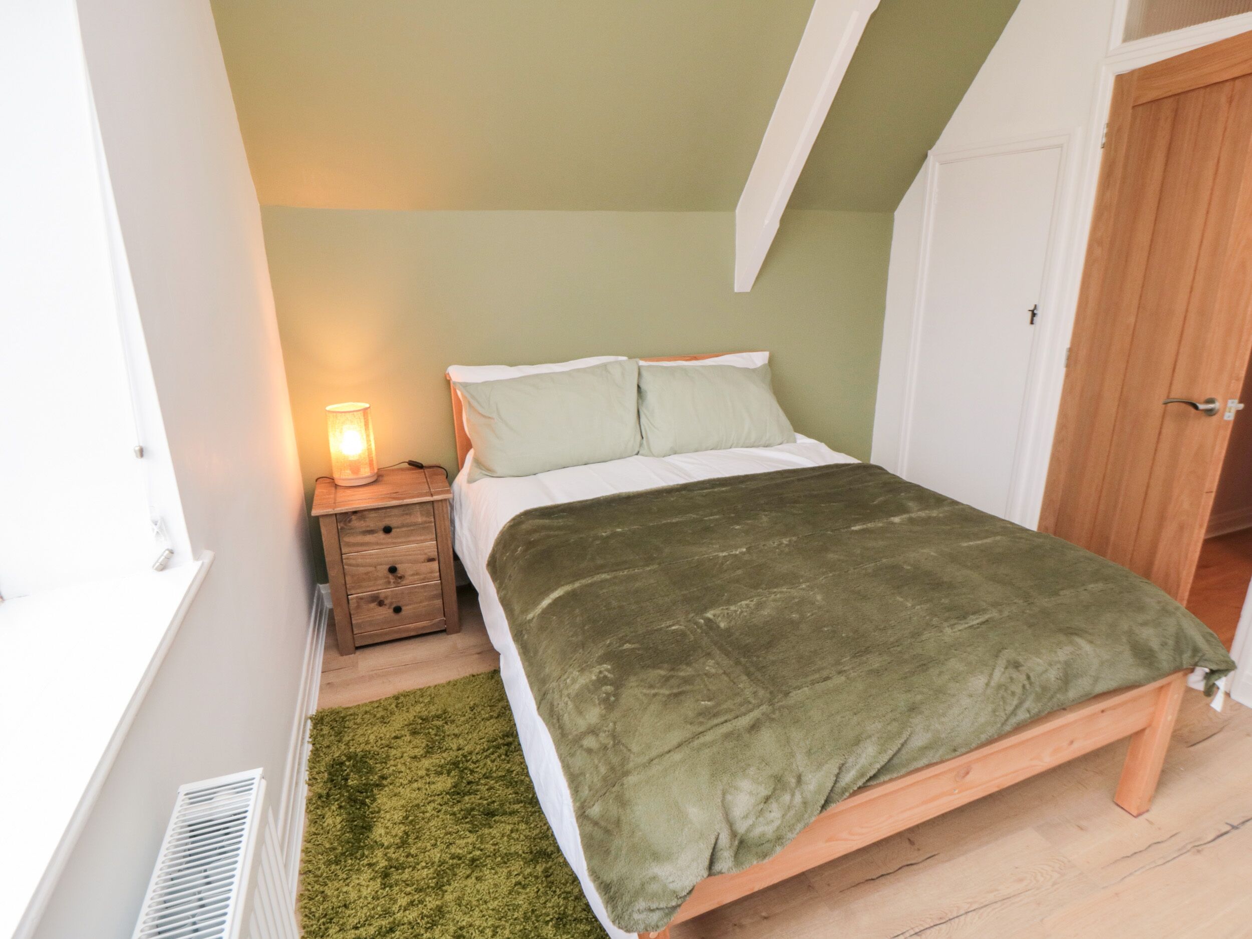 Une chambre à coucher avec un lit, une table de chevet avec une lampe, un tapis vert et une armoire en bois.