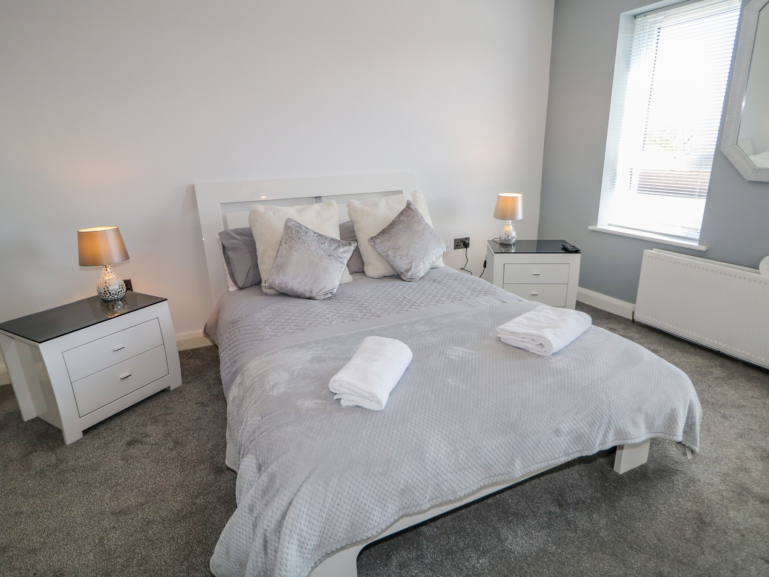 A neatly made bed with a grey quilt, two bedside tables with lamps, and a window with blinds.