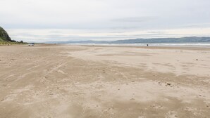 Ein weiter, offener Strand mit einigen Personen und einem Auto, und in der Ferne Hügel unter bewölktem Himmel.