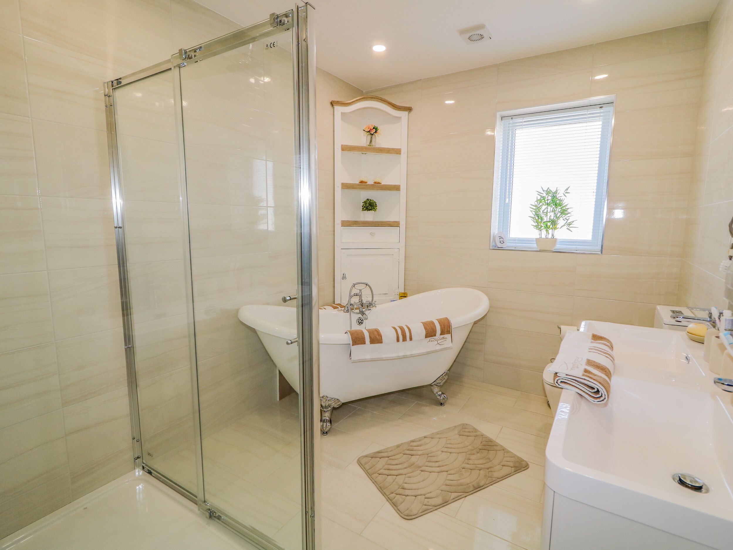 A bathroom with a glass-enclosed shower, a freestanding bathtub, a sink, and a window with a plant.