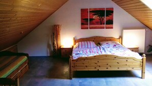 Ein Schlafzimmer mit schrägen Wänden, einem hölzernen Bett, einem Nachttisch, einem Beistelltisch, einem Stuhl und einem Bild an der Wand.