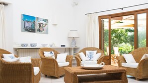 A room with wicker furniture, a white tiled floor, a table with a plate, and a window with a view of greenery.