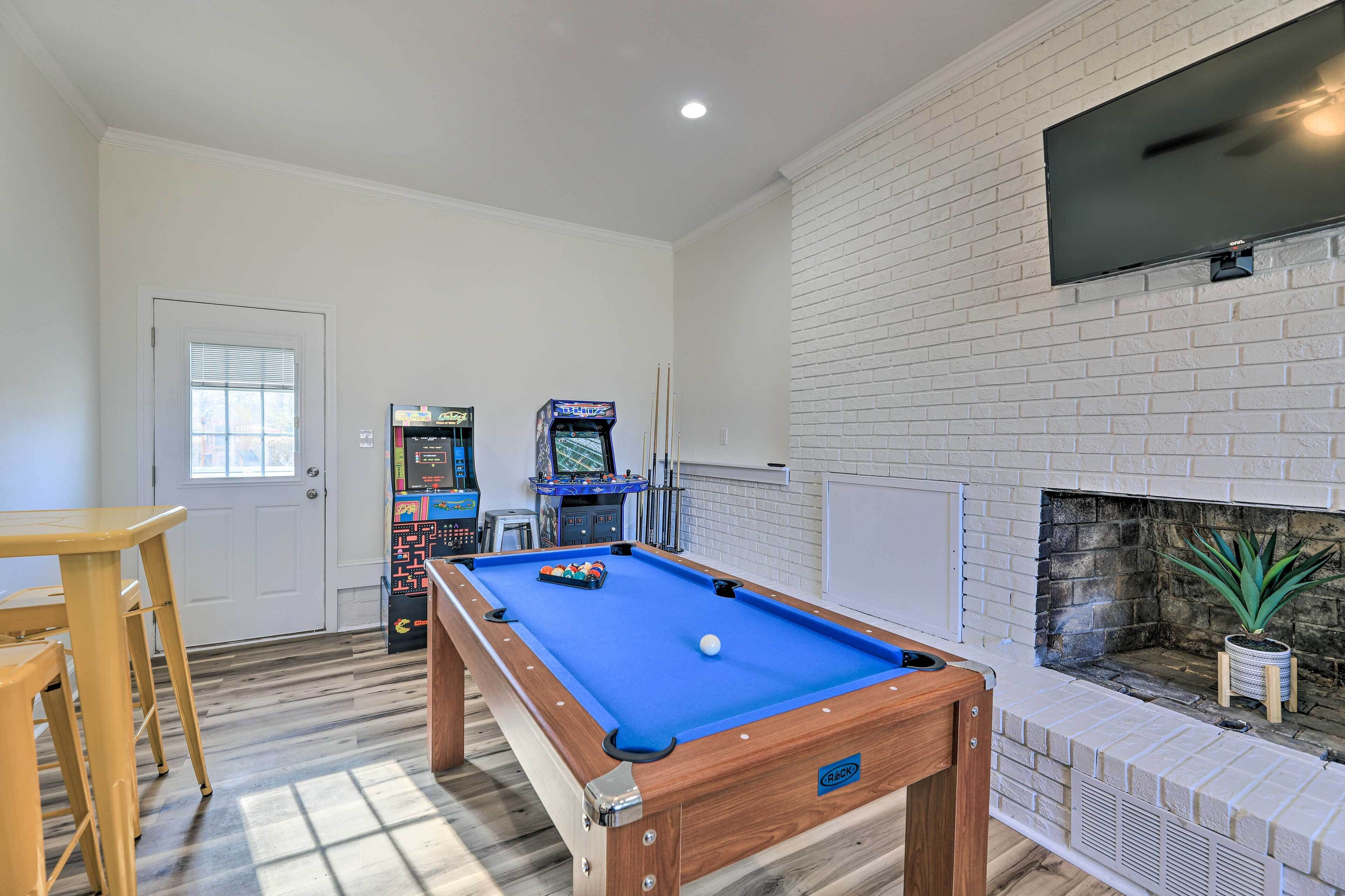 A pool table with a blue felt surface, a fireplace with a potted plant, and two arcade machines in the background.