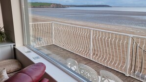 A room with a view of a beach, a window with a white metal railing, a red leather sofa, and a small table with a card on it. - Beach House (Duncannon)