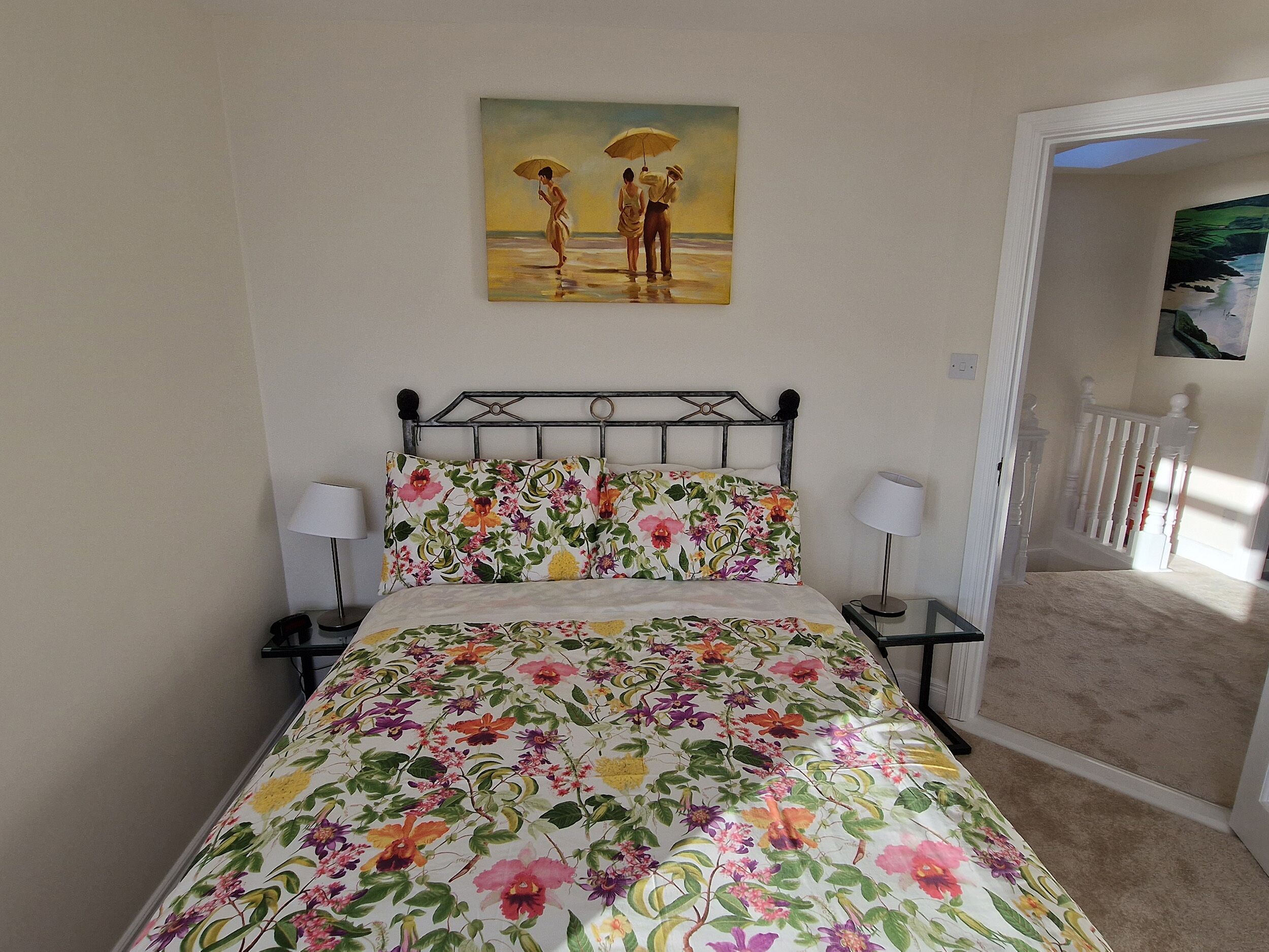 Ein Schlafzimmer mit floralem Bettbezug, zwei Nachttischlampen, einem Wandbild von zwei Personen und Blick auf einen Balkon.