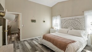 Quarto com cama grande, piso de madeira, mesa de cabeceira branca e cabeceira decorativa.