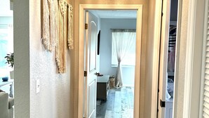 Um corredor com um candelabro, decoração de parede em macramé e vista para um quarto com janela e cortinas.