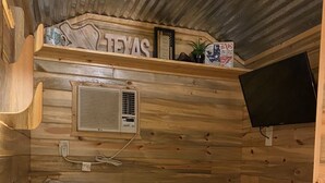 A cozy cabin room with a bed, pillows, a lamp, an air conditioning unit, and a mounted TV. - Cabin/Farmhouse w/fire pit and wide open spaces (Haslet)