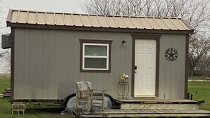 A small, single-story shed with a door and a window, a chair, and a small table outside. - Cabin/Farmhouse w/fire pit and wide open spaces (Haslet)