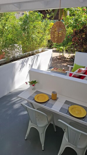An outdoor dining area with a table set for two, surrounded by white chairs, a woven basket hanging above, and a variety of plants in the background.