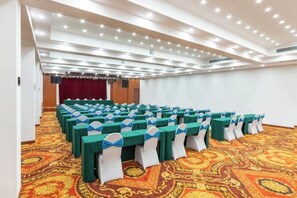 Un salón de conferencias con mesas dispuestas en filas, alfombra estampada y un escenario con cortinas.