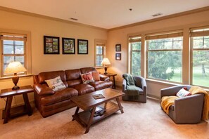 A living room with a brown leather sofa, a wooden coffee table, and two armchairs. There are framed pictures on the wall and a lamp on a side table.
