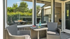 Une terrasse extérieure avec des chaises et une table en osier, une porte vitrée donnant sur une cuisine et une vue sur la verdure environnante.