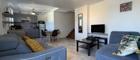 A modern living room with a sofa, a coffee table, and a TV. There is a kitchen area with cabinets and a microwave.