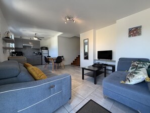 A modern living room with a sofa, a coffee table, and a TV. There is a kitchen area with cabinets and a microwave.