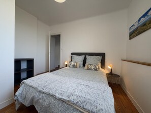 Ett sovrum med en stor säng, två sänglampor, ett nattduksbord, en trähylla och en dörr som leder till ett annat rum.
