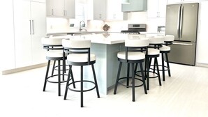 Une cuisine moderne avec des meubles blancs, un îlot central doté de tabourets, des appareils en acier inoxydable et un éclairage suspendu.