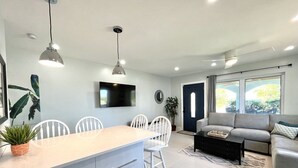 Un salon moderne avec un canapé gris, un coin repas avec des chaises blanches et une cuisine dotée d’un grand îlot central.