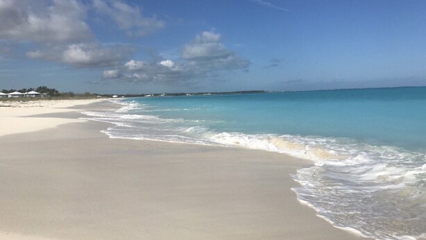 Une plage à l’eau turquoise et limpide, un rivage sablonneux et quelques bâtiments visibles au loin.