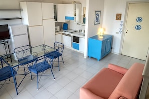 A compact kitchen with white cabinets, a microwave, and a blue chair. There is a glass table with blue chairs and a pink sofa in the living area.