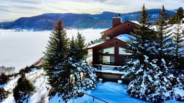 A cabin with a large window, surrounded by snow-covered trees and a lake.