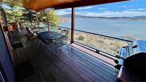 A wooden deck with a table and chairs, overlooking a body of water and distant hills.