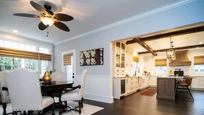 Un comedor amplio con una mesa de madera oscura y sillas blancas tapizadas, un ventilador de techo y vistas a la cocina contigua con armarios blancos y una isla grande.
