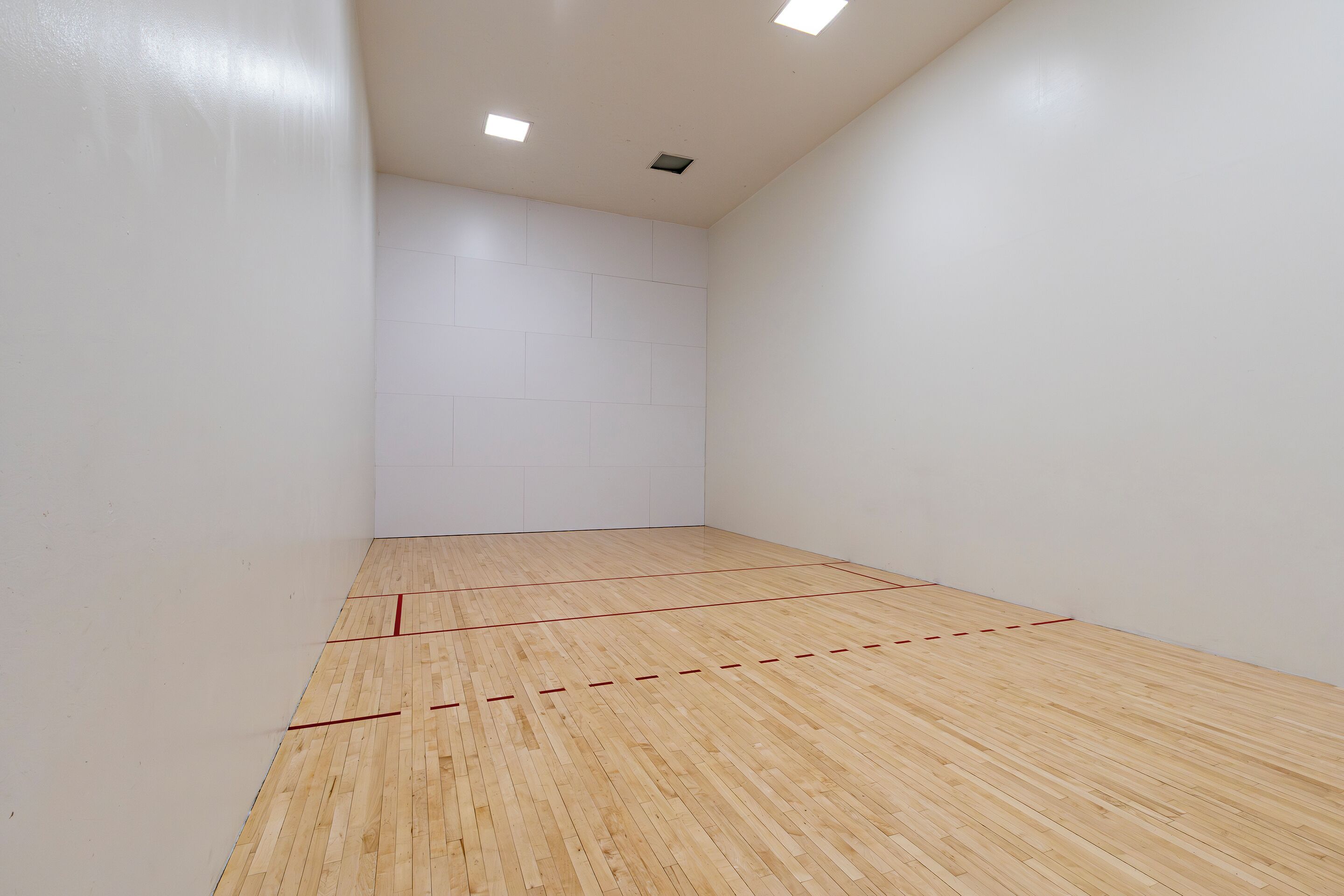 A squash court with white walls and a wooden floor marked with red lines.
