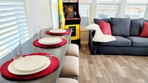 En matplats med ett långt bord som är dukat för fyra personer, en soffa och en bokhylla med en spelkonsol.