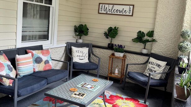 A cozy porch with a blue sofa, a glass coffee table, and a floral rug. There are two chairs with cushions, a 'Welcome' sign, and a 'Stay awhile' pillow. - Charming Victorian at Lake Shelbyville ⋅ Sleeps 6 ⋅Sunroom ⋅ Fire Pit ⋅ Parking (Shelbyville)